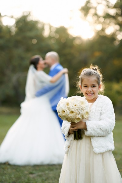 Fotka dcerky mladomanzelov stojaca vpredu so svadobnou kyticou, vzadu bozkavajuci sa rodicia